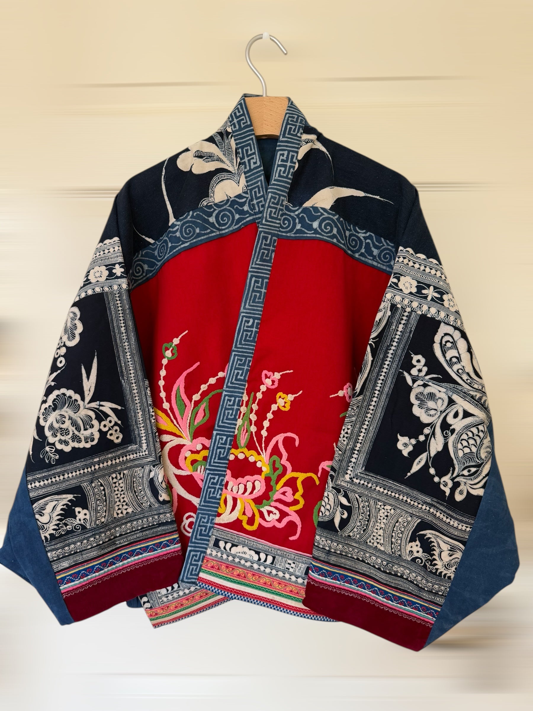 Traditional jacket with red and blue patterns on a wooden hanger against a beige wall.